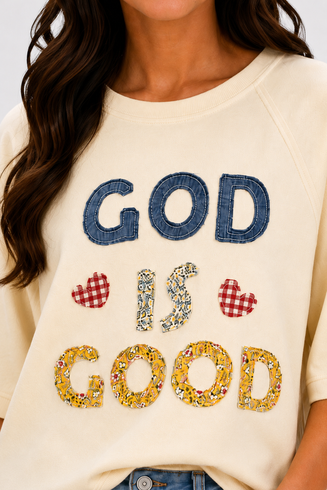 A close-up of a woman wearing a cream-colored sweatshirt with a decorative graphic that reads 'GOD IS GOOD' in blue denim, floral print, and red-and-white gingham hearts.