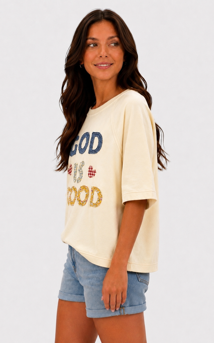 A woman modeling an oversized cream-colored graphic t-shirt with 'GOD IS GOOD' printed on the front in blue, red, and gold glitter-style lettering, paired with light blue denim shorts against a plain white background.