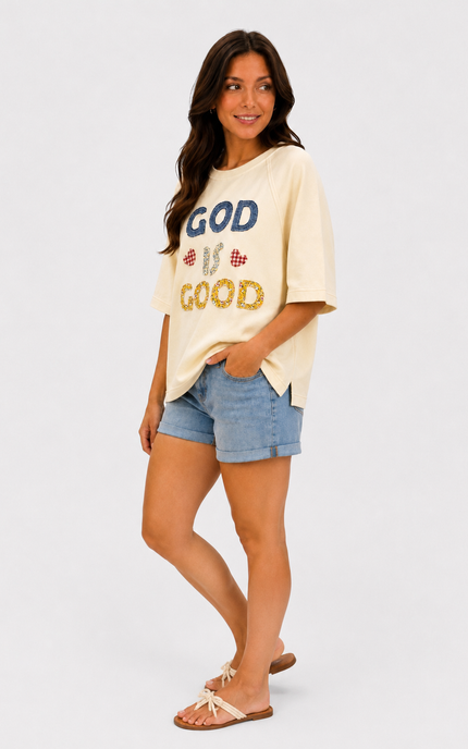 A woman modeling a cream-colored graphic t-shirt with 'GOD IS GOOD' text in blue and gold sequins, paired with denim shorts and flat sandals, against a light gray studio background.