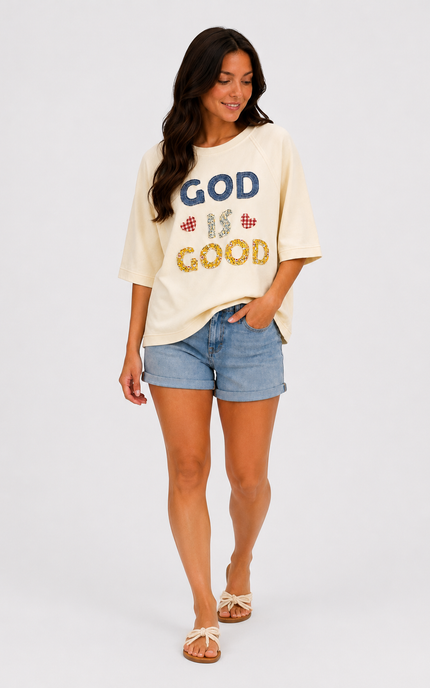 A woman modeling a cream-colored graphic t-shirt with 'GOD IS GOOD' written on the front in blue and gold glitter-style lettering, paired with denim shorts and beige sandals, against a light gray studio background.