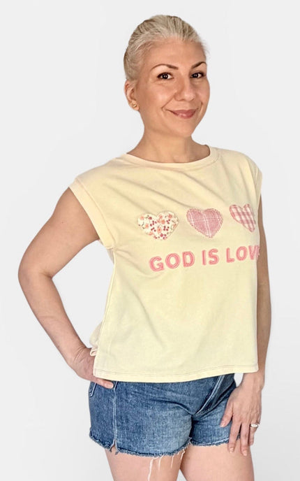 A woman modeling a pale yellow sleeveless crop top with pink heart graphics and the text 'GOD IS LOVE' printed on the front, paired with denim shorts against a light gray background.