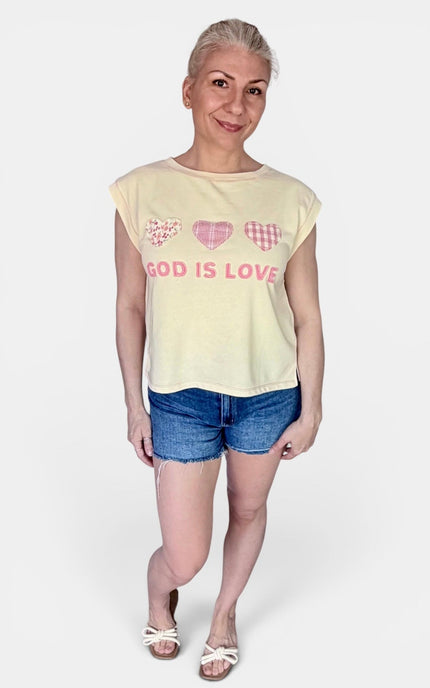 A woman modeling a pale yellow sleeveless t-shirt with pink heart graphics and the text 'GOD IS LOVE' on the front, paired with denim shorts and white sandals, against a light gray background.