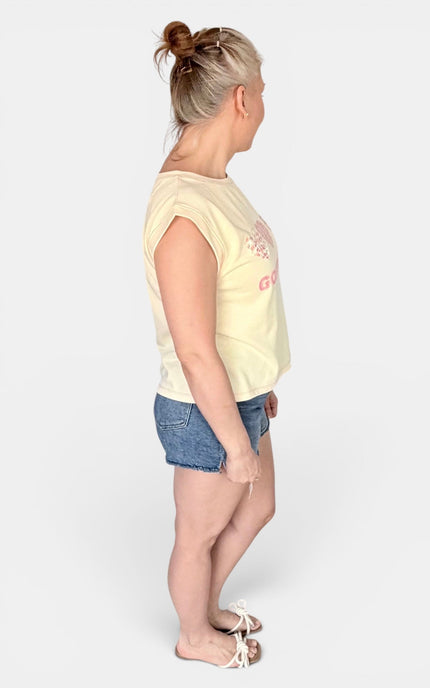 A woman shown from the side profile wearing a pale yellow sleeveless top with a pink graphic print, blue denim shorts, and white sandals, standing against a light gray studio background.