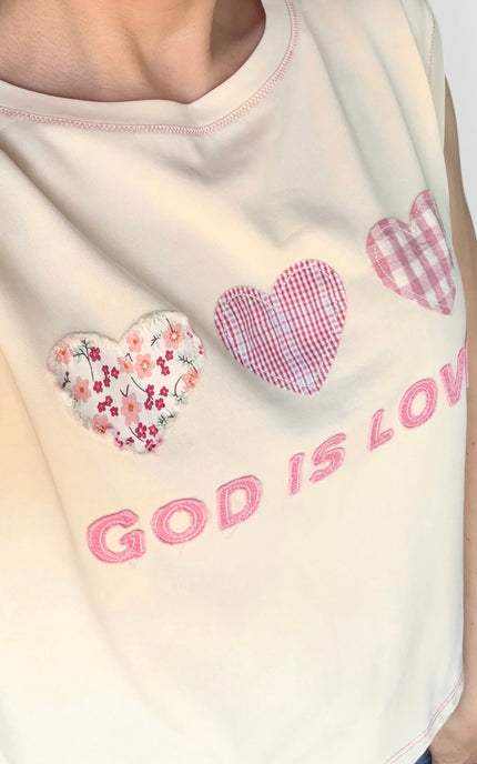 A close-up of a cream-colored sleeveless top featuring three decorative hearts in different patterns (floral, pink-and-white gingham, and pink-and-white check) with the embroidered pink text 'GOD IS LOVE' below them.