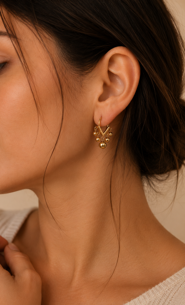 Woman wearing open V-shaped 18k gold hoop earrings with polished dangling ball beads for a modern, elegant look.