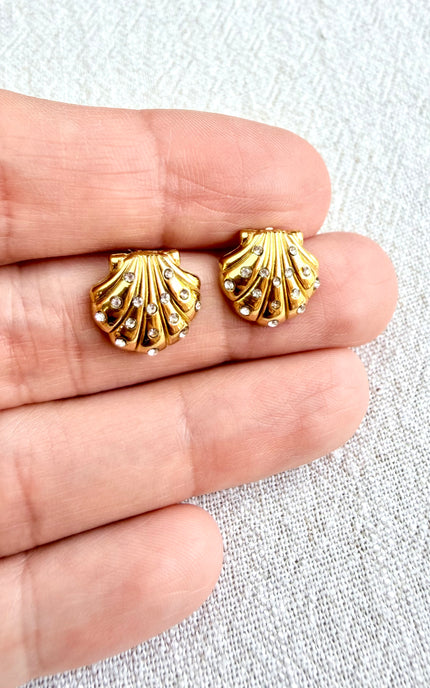 Gold shell-shaped earrings held between fingers on a light gray background