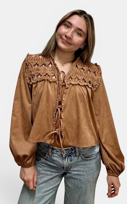 Cropped tan suede-like blouse with a large crocheted lace yoke and tie-front detailing, paired with light-wash blue jeans, against a plain light gray studio background.