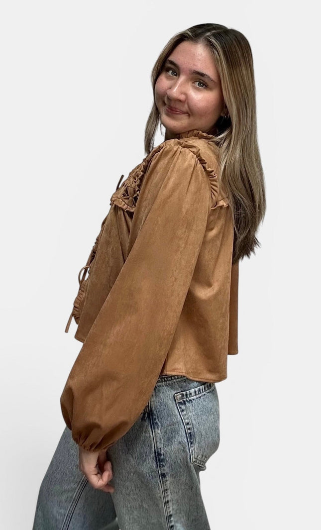 Cropped tan suede-like blouse with lace-up details and ruffled trim, paired with light-wash jeans, photographed against a plain light gray studio background.