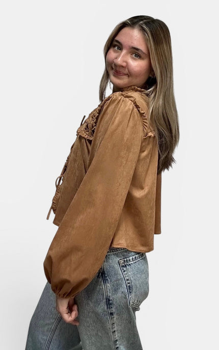 Cropped tan suede-like blouse with lace-up details and ruffled trim, paired with light-wash jeans, photographed against a plain light gray studio background.