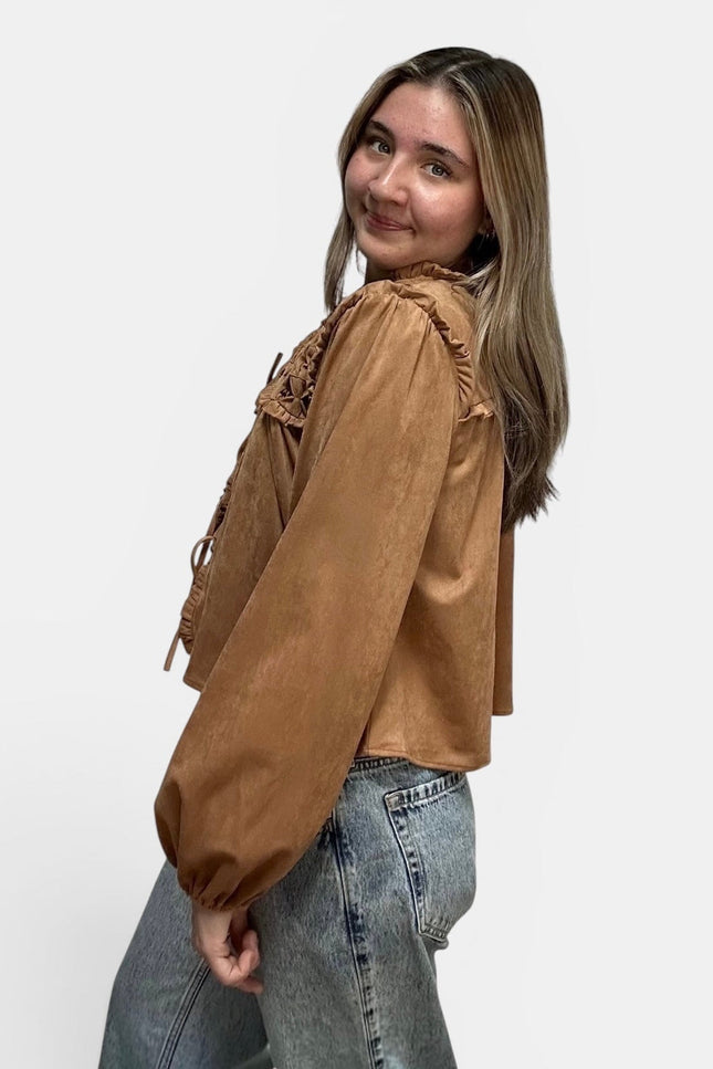 Cropped tan suede-like blouse with lace-up details and ruffled trim, paired with light-wash jeans, photographed against a plain light gray studio background.
