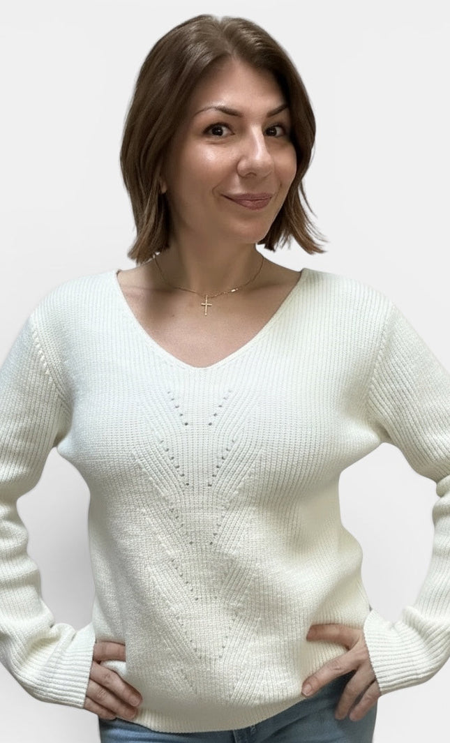 A woman with short brown hair wearing a cream-colored ribbed knit sweater with a V-neck and delicate knit pattern on the chest, paired with blue jeans, against a plain light gray background.