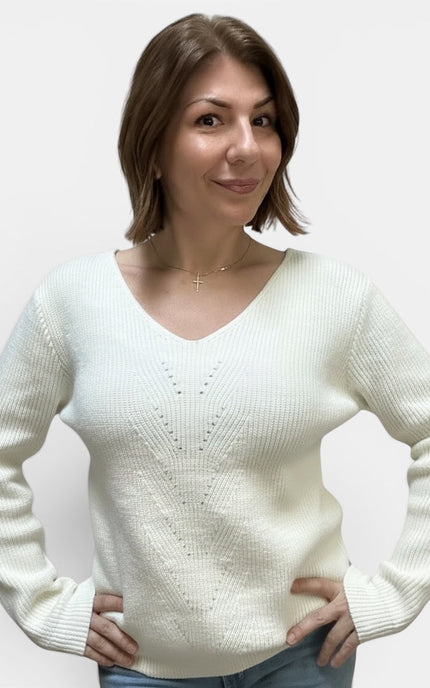 A woman with short brown hair wearing a cream-colored ribbed knit sweater with a V-neck and delicate knit pattern on the chest, paired with blue jeans, against a plain light gray background.