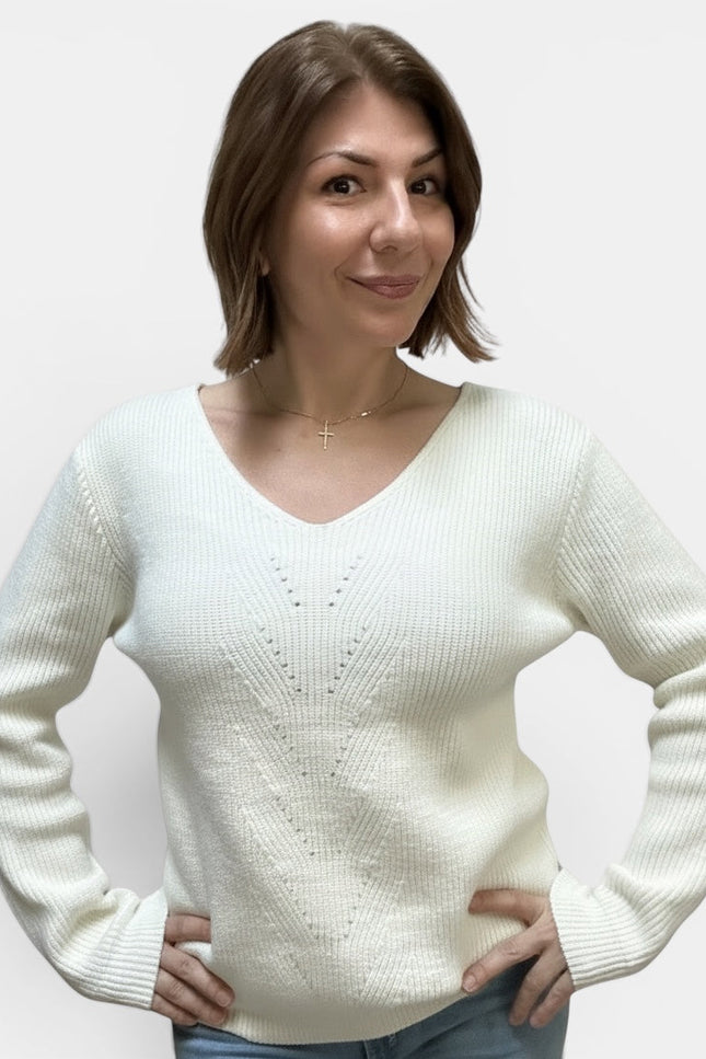 A woman with short brown hair wearing a cream-colored ribbed knit sweater with a V-neck and delicate knit pattern on the chest, paired with blue jeans, against a plain light gray background.