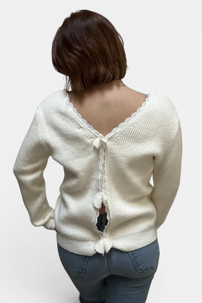 A woman viewed from behind wearing a cream-colored ribbed knit cardigan with a lace-trimmed open back and decorative tie details, paired with blue jeans, against a plain light gray background.