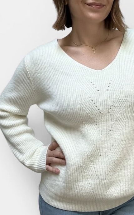 A woman wearing a ribbed knit V-neck sweater in off-white, paired with blue jeans and a delicate gold necklace, against a plain light background.