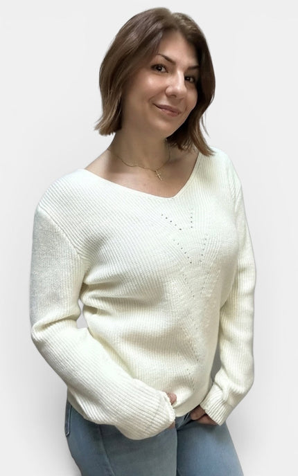 A woman wearing a cream-colored, long-sleeve knit sweater with a V-neck and textured ribbing, paired with blue jeans, against a plain light gray background.