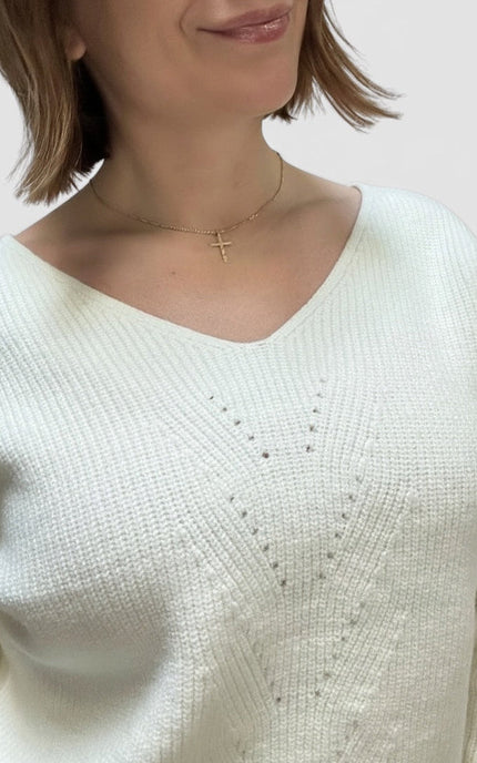 A close-up of a woman wearing a cream-colored knit sweater with a delicate gold cross necklace, set against a plain light background.