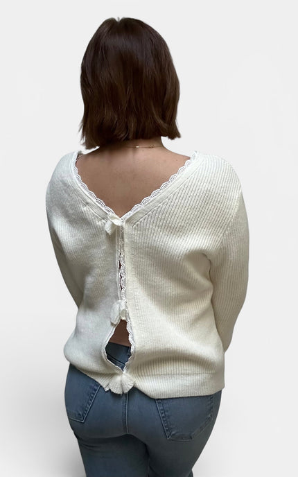 A woman viewed from the back wearing a cream-colored ribbed knit sweater with a lace-trimmed open back and tie closure, paired with blue jeans, against a plain light gray background.
