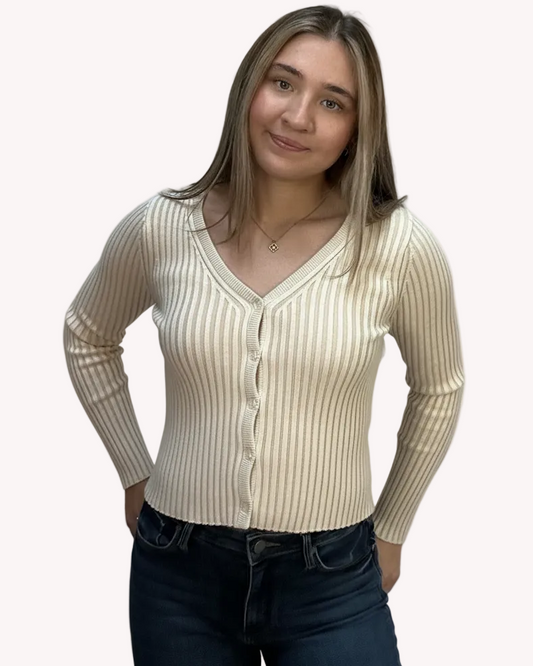 Woman wearing a beige ribbed cardigan over a white shirt with dark jeans on a light pink background