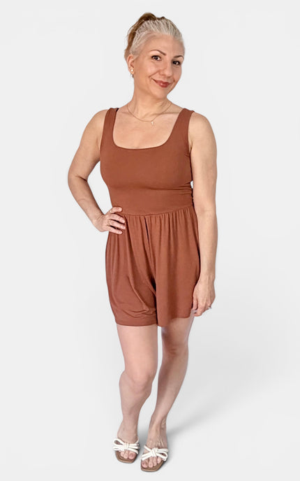 A woman modeling a rust-brown sleeveless romper with a scoop neckline and short, gathered legs, standing against a light gray studio background.