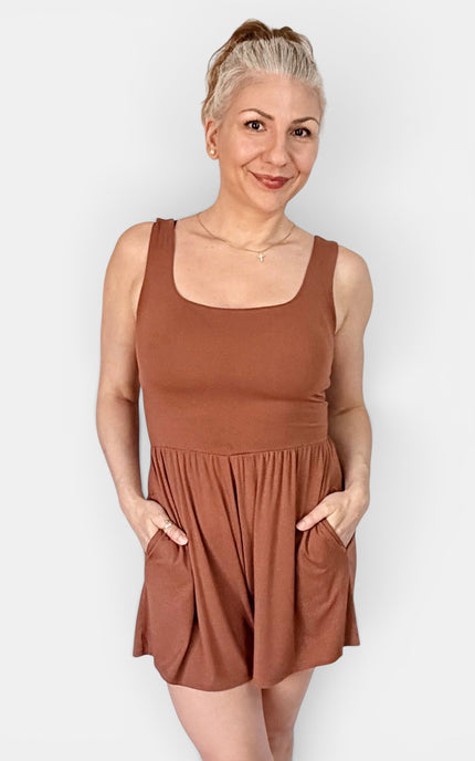 A woman modeling a sleeveless rust-brown romper with a scoop neckline and side pockets, standing against a light gray studio background.