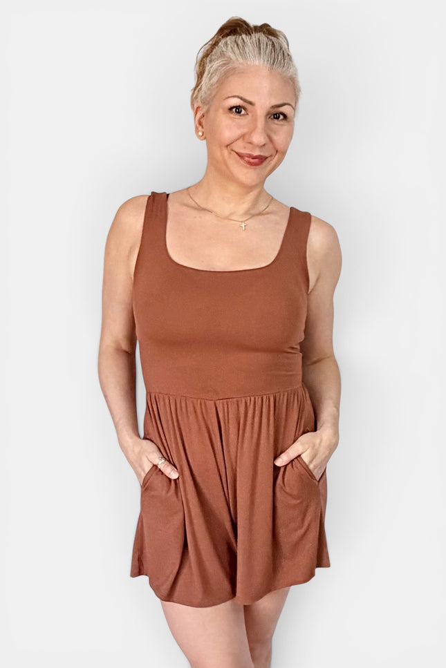 A woman modeling a sleeveless rust-brown romper with a scoop neckline and side pockets, standing against a light gray studio background.