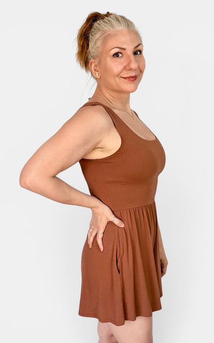 A woman modeling a sleeveless, scoop-neck mini dress in a warm rust brown color, standing against a light gray studio background.