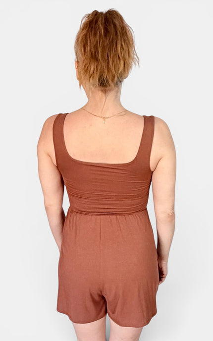A woman viewed from the back wearing a rust-brown sleeveless romper with a square neckline and short legs, against a light gray studio background.