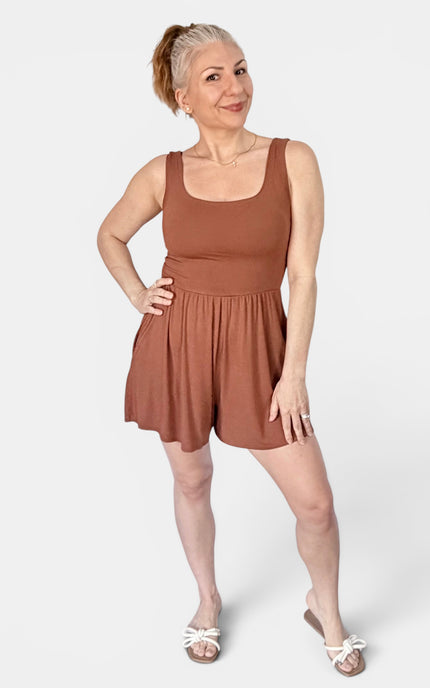 A woman modeling a sleeveless rust-brown romper with a scoop neckline and flared shorts, standing against a light gray studio background.