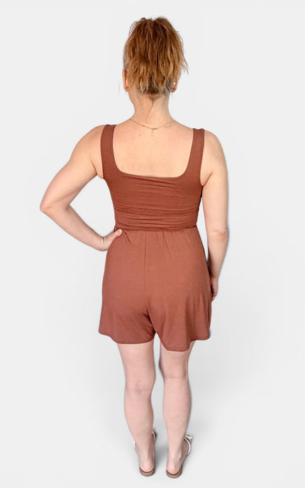 A woman viewed from the back wearing a rust-brown sleeveless romper with a square neckline and short legs, standing against a light gray studio background.