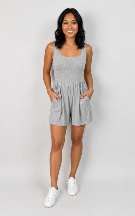 A woman modeling a light heather gray sleeveless romper with side pockets, paired with white sneakers, against a light gray studio background.