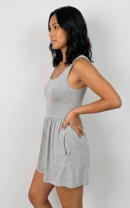 A woman modeling a light gray heather sleeveless mini dress with a gathered waist and side pockets, posed against a plain light gray background.