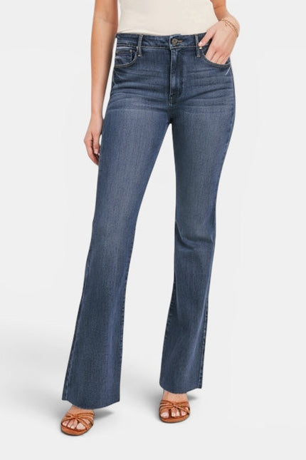 A model wearing a pair of medium-wash blue flare jeans with side slits, paired with a cream top and brown sandals, shown from the waist down on a light gray background.
