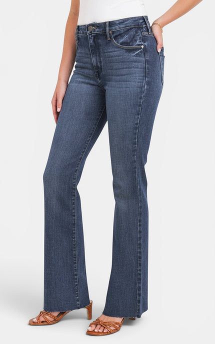 A model wearing a pair of medium-wash blue flare jeans with a white top and brown heeled sandals, shown from the waist down against a light gray studio background.