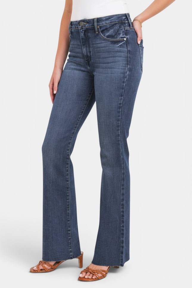 A model wearing a pair of medium-wash blue flare jeans with a white top and brown heeled sandals, shown from the waist down against a light gray studio background.