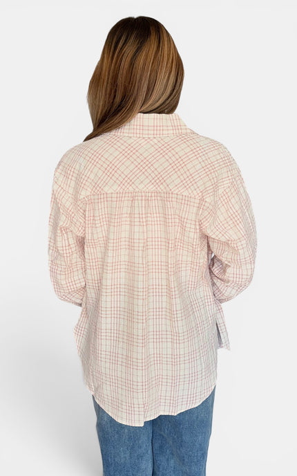 A woman viewed from the back wearing a light pink and white plaid button-up shirt with blue jeans, set against a plain light gray background.