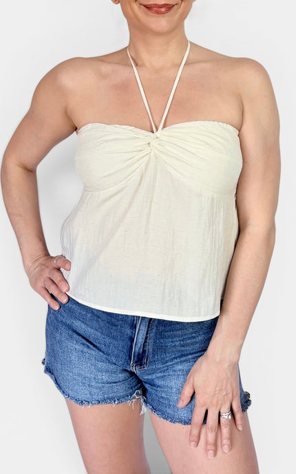 A woman wearing a cream-colored halter strap top with a gathered sweetheart neckline and blue denim shorts, shown from the chin down against a light gray background.