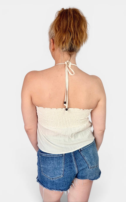A woman viewed from the back wearing a cream-colored halter strap top and denim shorts against a plain light gray background.