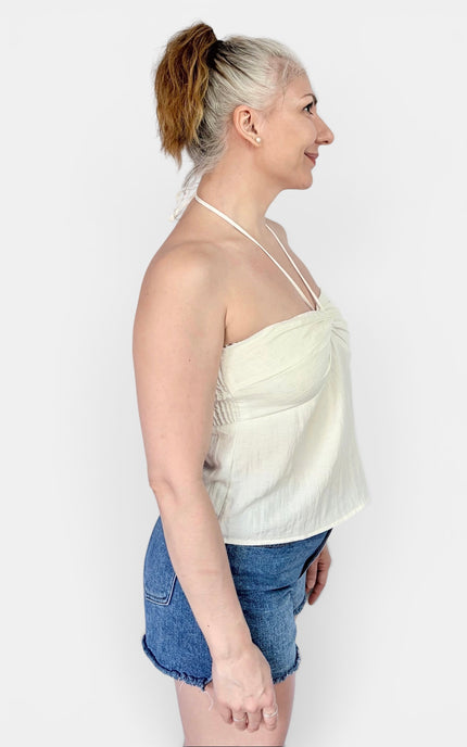 A woman shown in side profile wearing a cream-colored halter crop top and denim shorts against a plain light gray background.