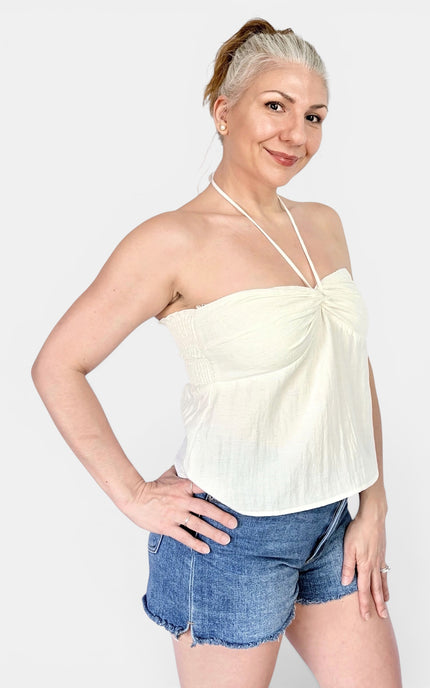 A woman modeling a cream-colored halter neck top with a twisted front detail, paired with denim shorts, against a light gray studio background.