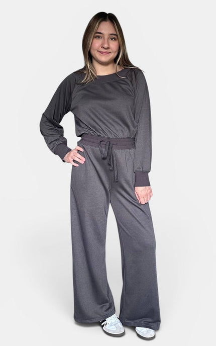 A woman modeling a dark gray two-piece loungewear set consisting of a long-sleeve crewneck top and matching wide-leg pants with a drawstring waist, against a plain light gray background.