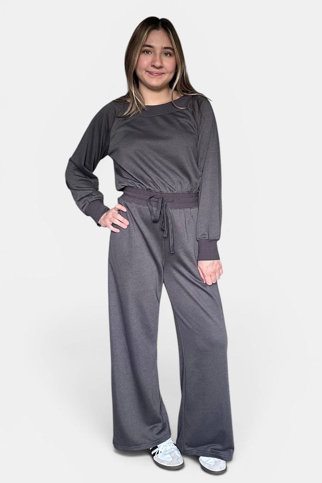 A woman modeling a dark gray two-piece loungewear set consisting of a long-sleeve crewneck top and matching wide-leg pants with a drawstring waist, against a plain light gray background.