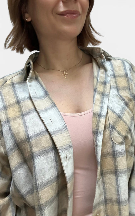 A close-up of a person wearing an oversized plaid button-up shirt in beige, cream, and gray tones over a light pink tank top, with a delicate gold cross necklace visible.