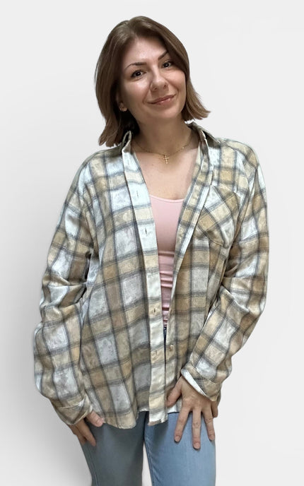 A woman modeling a long-sleeve plaid button-up shirt in beige, white, and gray tones, worn open over a light pink top and blue jeans, against a plain light gray background.