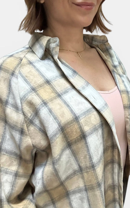 A close-up of a person wearing a light beige and white plaid button-up shirt over a light pink top, with a thin gold necklace visible against a plain light background.