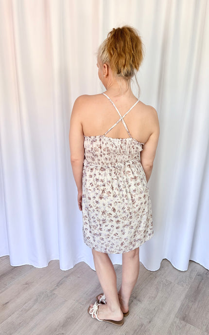 Back angled view of a lavender floral mini dress featuring a smocked back, tie detail, and pockets.