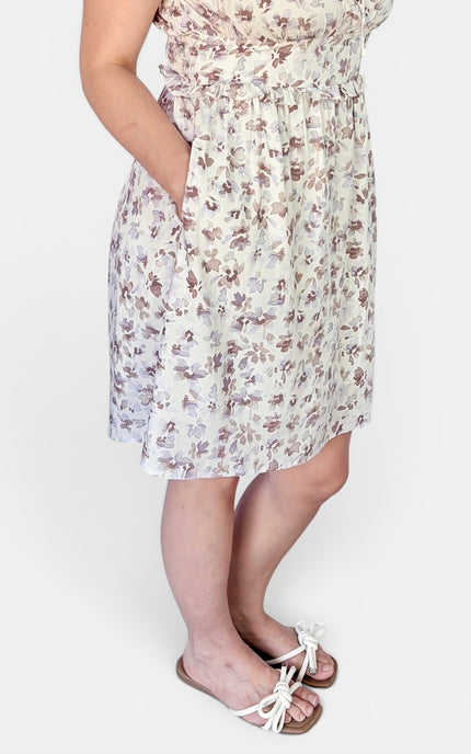 A woman wearing a sleeveless knee-length dress with a small floral print in muted brown and beige tones on a light cream background. The dress has side pockets and a gathered waist.