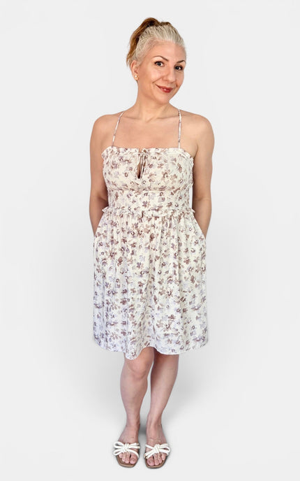 A woman modeling a sleeveless spaghetti-strap mini dress with a small brown and beige floral print on a white background, paired with white slide sandals, against a light gray studio background.