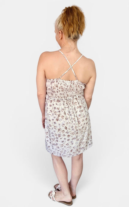 A woman viewed from behind wearing a sleeveless white mini dress with a small brown floral print and thin crisscross back straps, paired with flat sandals, against a light gray studio background.