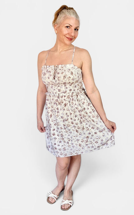 A woman modeling a sleeveless white spaghetti-strap mini dress with a small brown floral print, paired with white slide sandals, against a light gray studio background.
